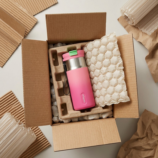 A top-down view shows the Contigo 16 oz Tween Dual Sip travel mug, in its pink and stainless steel ombre design, resting safely amidst recycled paper void-fill inside a sturdy shipping box. A white bubble-wrap bag is partially peeled away. The 'contigo' logo and all original product details are clearly visible on the item. The scene inspires confidence of safe delivery.