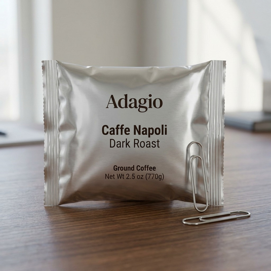 A single Adagio Caffe Napoli coffee packet stands next to standard-size paperclips on a wooden desk.
