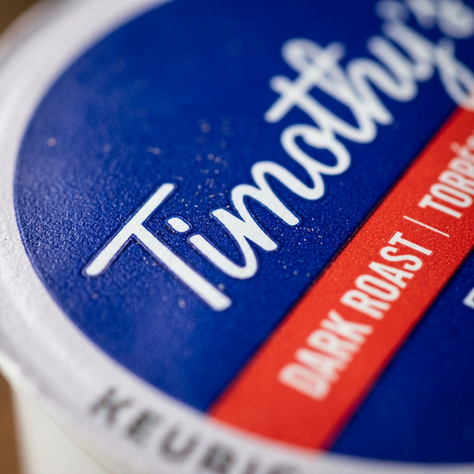 A macro photograph detailing the precise, sharp, white Timothy's coffee logo print on the deep blue K-Cup lid, showcasing the high-quality, professional manufacturing standards suitable for a Keurig brewer.