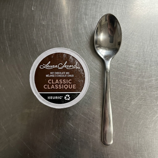 A top-down view of a Laura Secord Hot Chocolate K-Cup, exactly as seen in [image_0.png], placed neatly on a kitchen counter next to a common stainless steel teaspoon for scale. The lighting is soft and even.